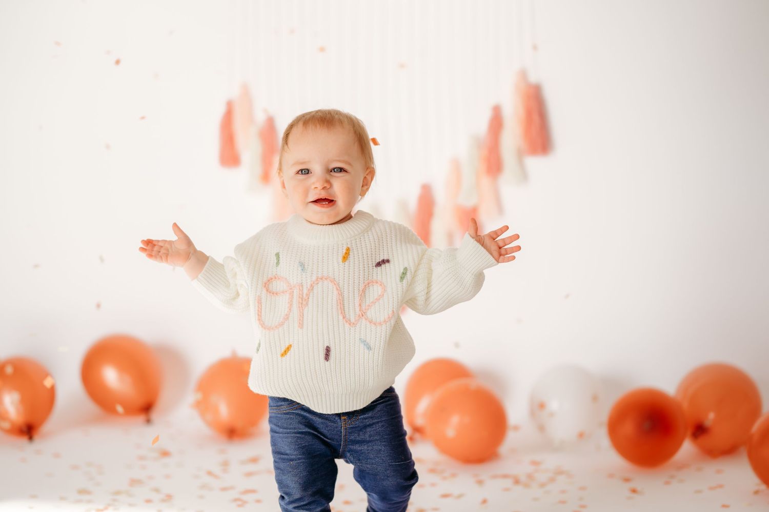 Small child in white sweater with coral decorations and balloons for birthday celebration.