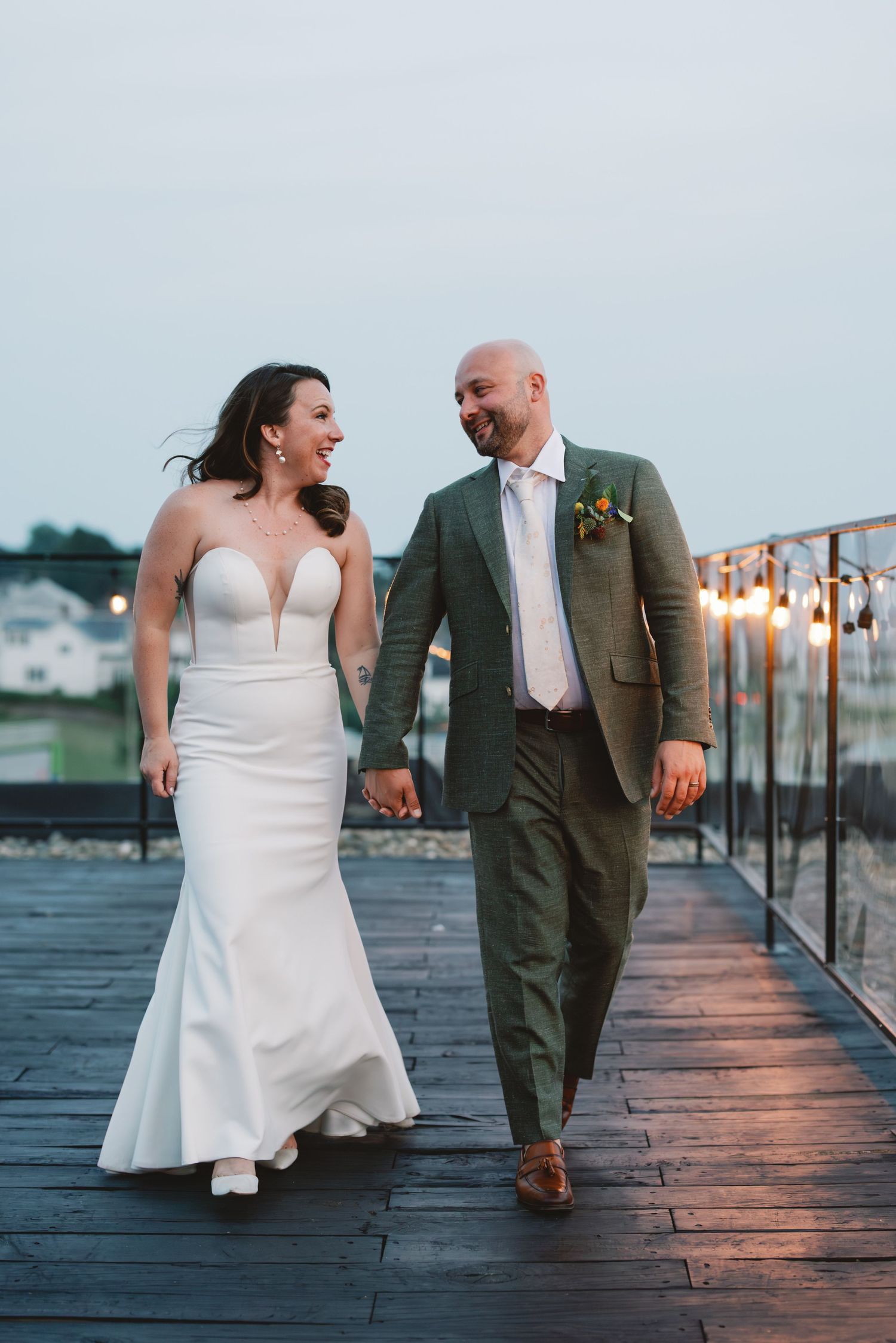 A couple shares a joyful moment walking hand in hand on a wooden deck during a sunset wedding celebration.