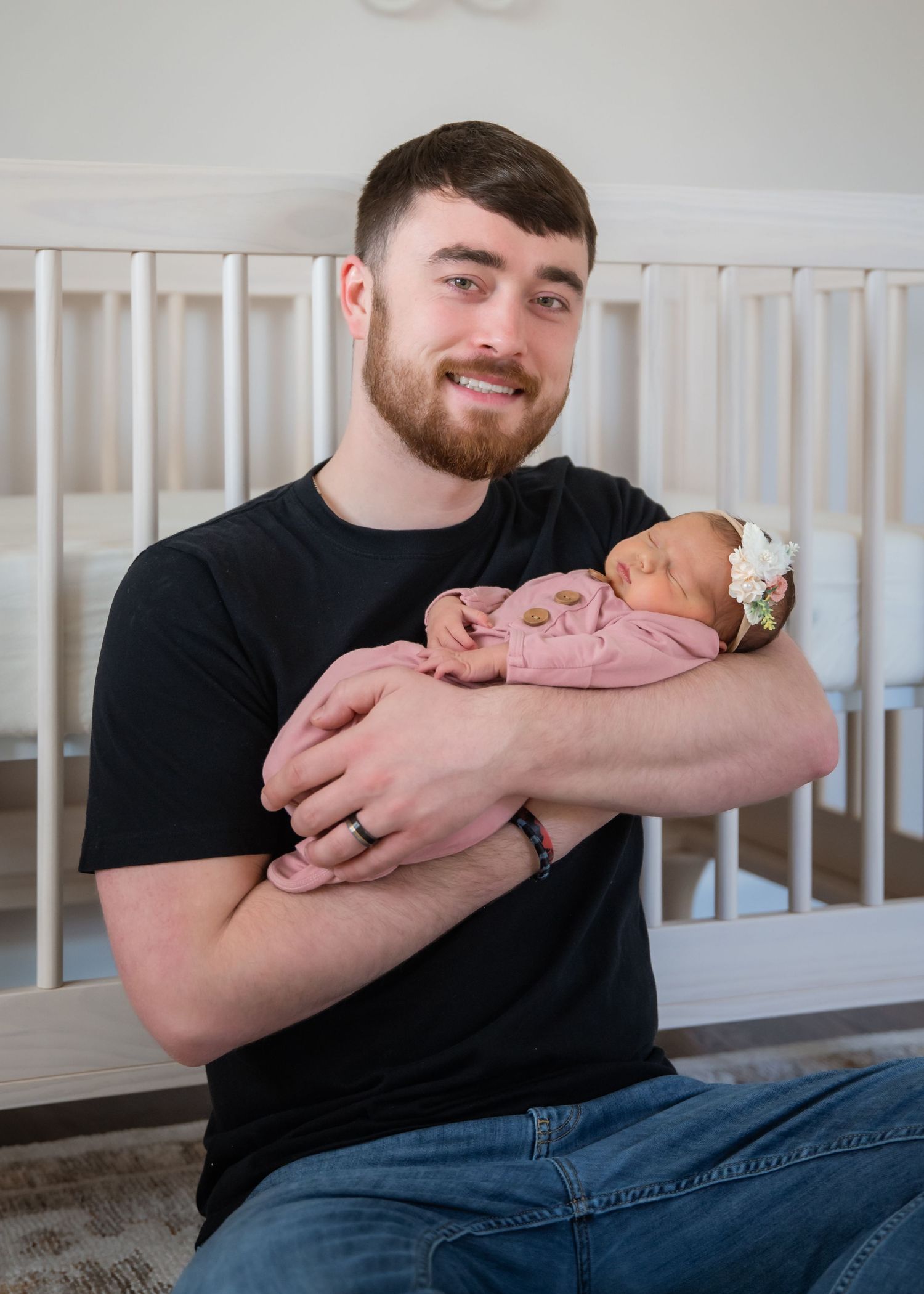 A tender moment shows someone in a black t-shirt holding a sleeping newborn baby in pink against a white crib background.