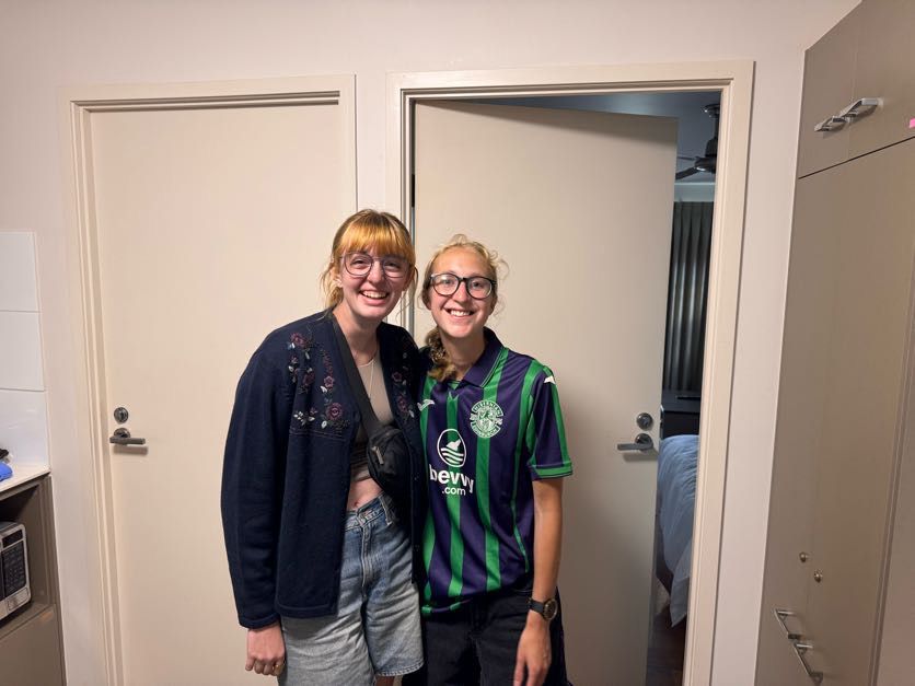 Two people posing together in a white-walled apartment hallway.