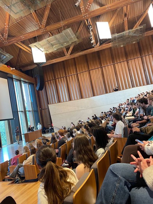 Large lecture hall filled with students during a presentation with wooden ceiling beams.