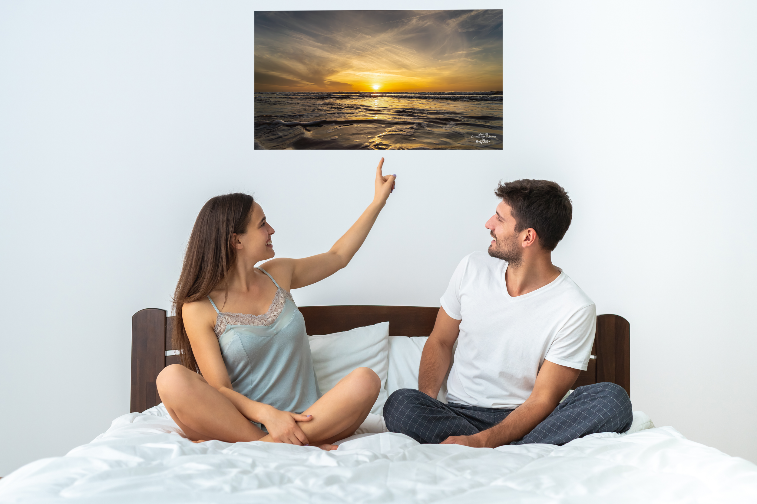 Two people sit on a bed while playfully interacting beneath a sunrise beach canvas wall art print.
