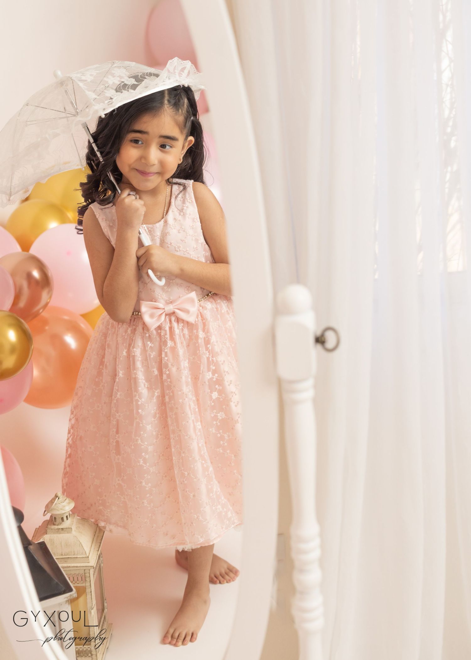 A child in a peach lace dress and white veil poses barefoot next to colorful balloons against a light curtained backdrop.