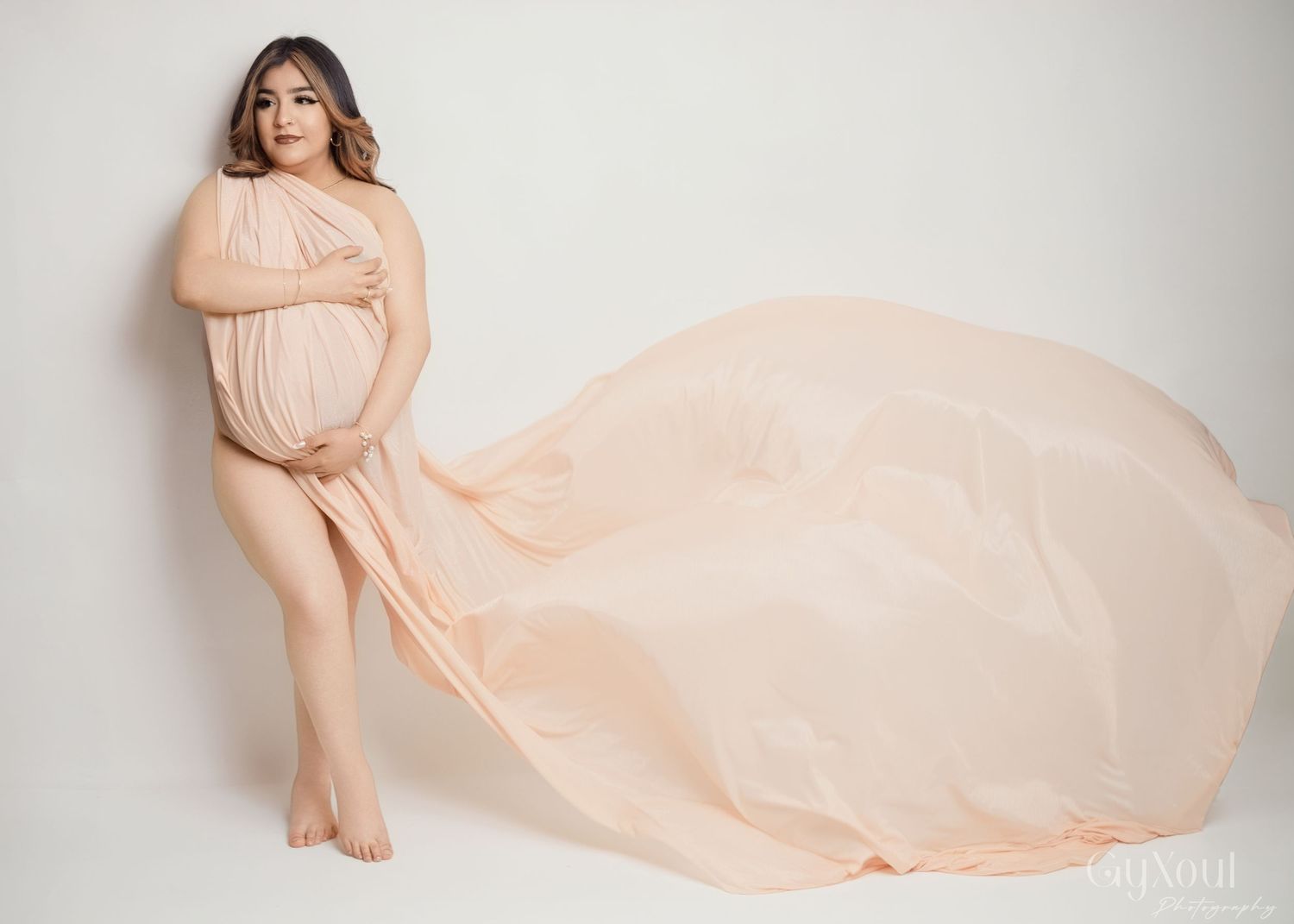 A flowing peach maternity gown billows dramatically against a light backdrop in an elegant studio portrait.