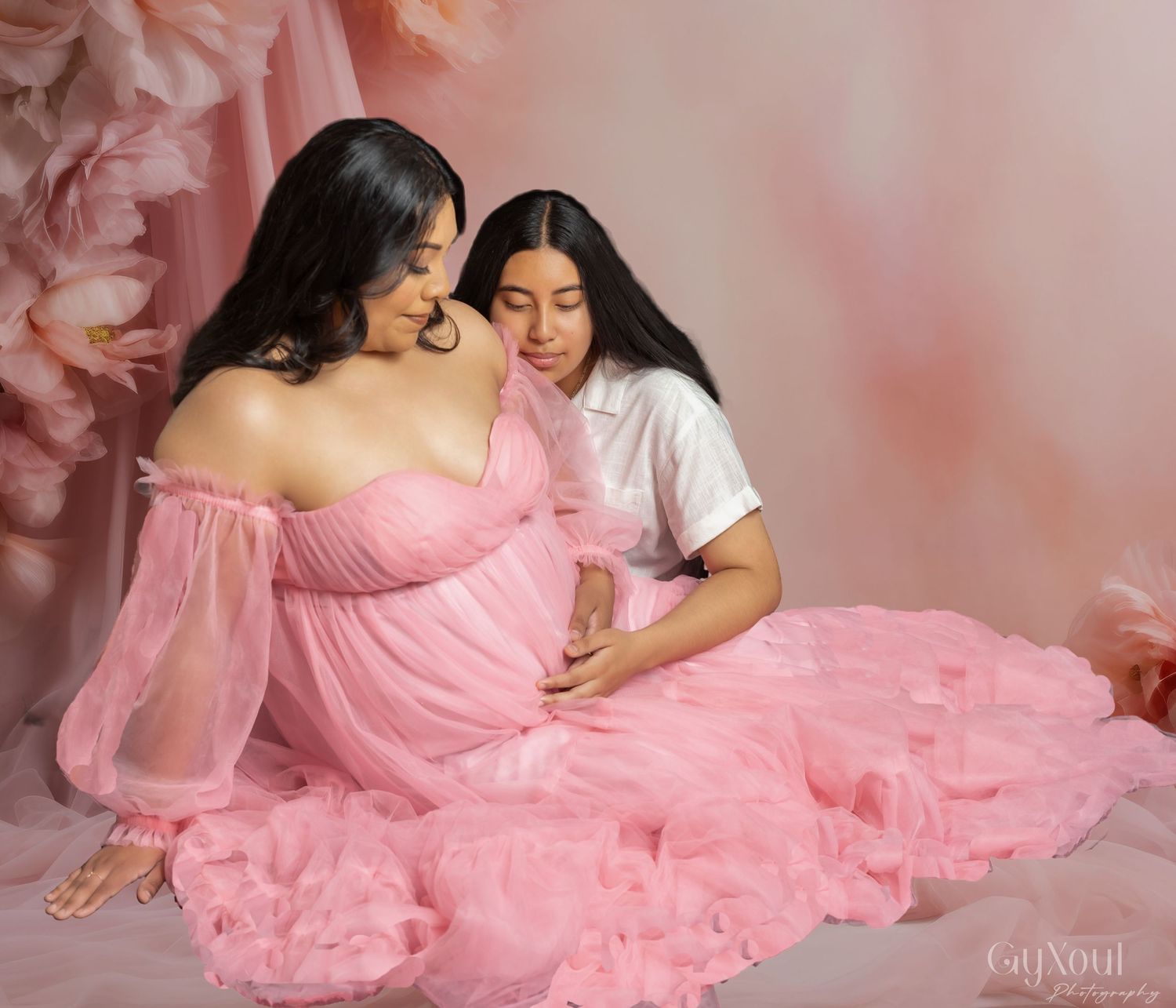 Two people in pink maternity photo setting with floral backdrop and flowing pink tulle dress.