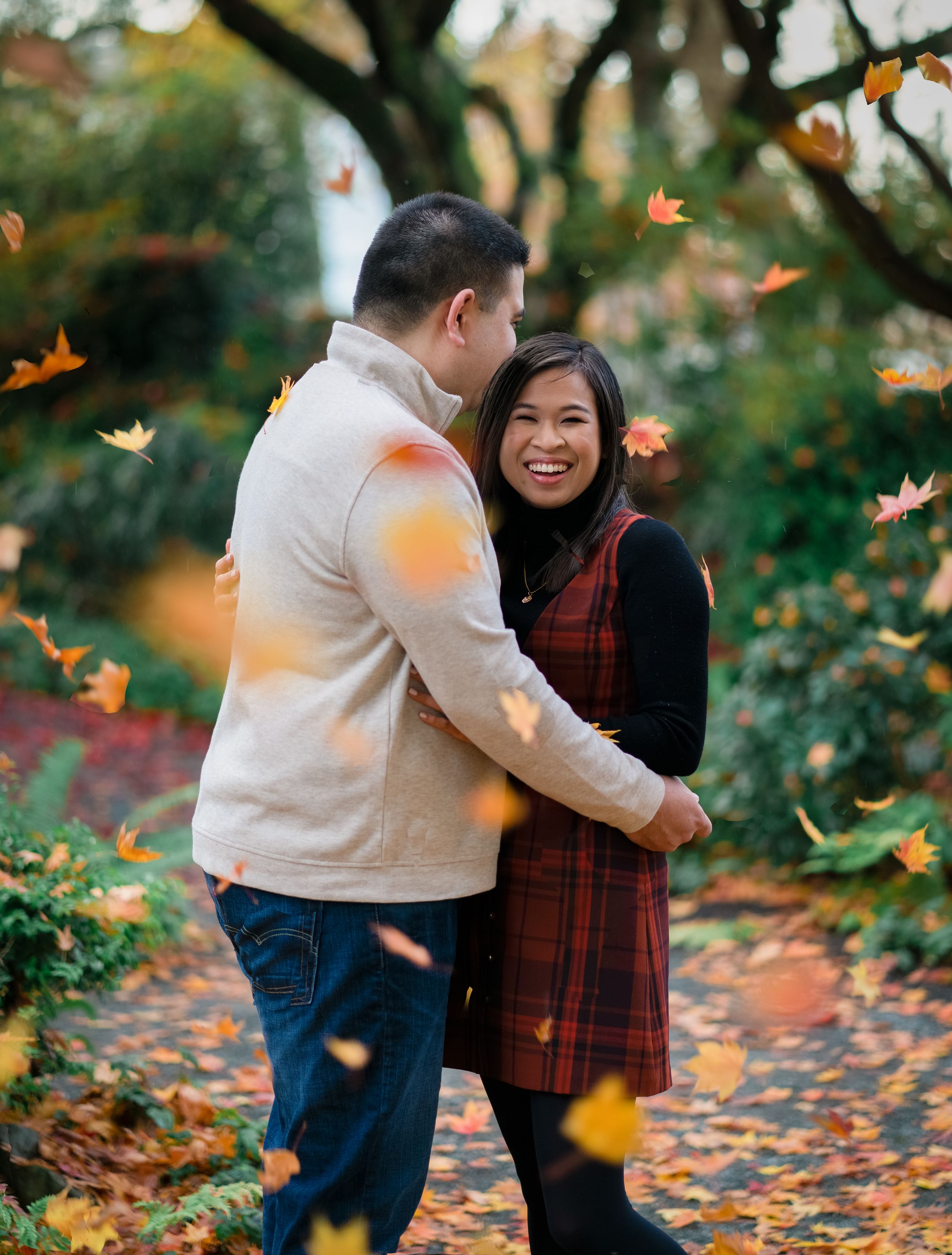 A couple enjoys a playful autumn photoshoot among falling orange leaves in a park.