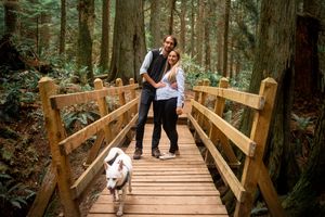 A couple stands on a wooden bridge in a forest with their dog.