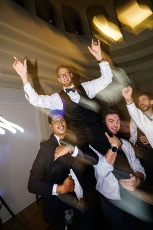 Groomsmen dance enthusiastically at a wedding reception under indoor lighting.