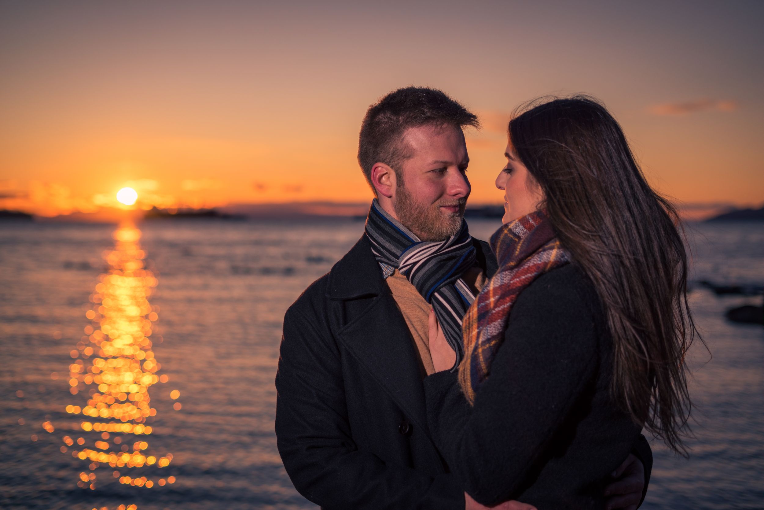 A romantic couple embraces on a beach at sunset with golden light reflecting on the ocean.