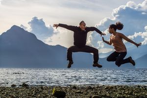 Two silhouettes jump joyfully against a backdrop of mountains and water during sunset.