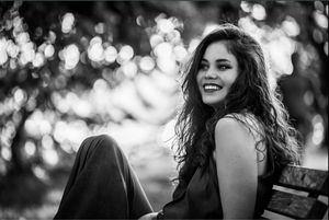 Black and white portrait of someone with long wavy hair sitting on a bench with a dreamy bokeh background.