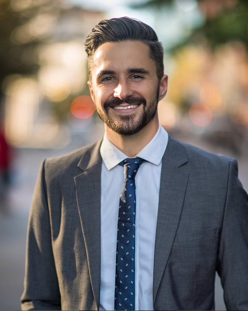 Professional headshot of someone smiling warmly while wearing a grey suit with white shirt and patterned navy tie.