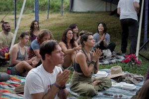 Festival attendees sit on blankets watching outdoor performance with tents in background.