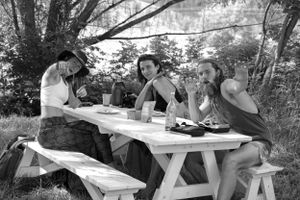 Friends share conversation and food at a picnic table in dappled sunlight.