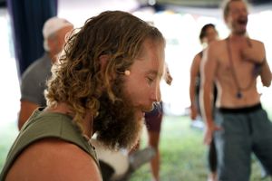 A bearded person in a green sleeveless shirt standing among others at an outdoor gathering.