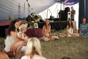 Group of people sitting on grass inside a tent with musical equipment in background.