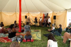 Musicians perform on stage while audience members sit on colorful mats in a festival tent.