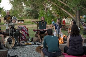 Musicians performing around a campfire at an outdoor gathering with audience sitting on the ground.