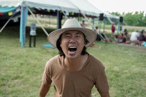 Person in a brown t-shirt and sun hat expressing joy at an outdoor festival.