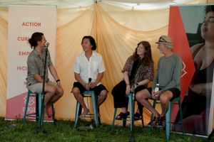 A group sits on stools having a discussion panel in front of a beige backdrop at an outdoor event.