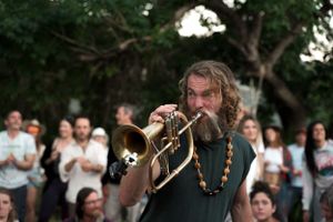 A bearded musician plays trumpet for an outdoor audience.