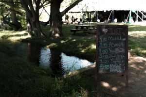 A chalkboard menu sign stands near a tranquil pond under a large shady tree.