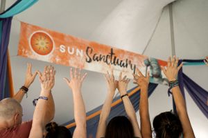 A group of people raise their hands toward a 'Sun Sanctuary' banner during an event.