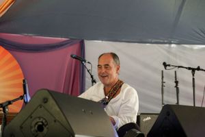 A performer plays at an outdoor festival event behind audio equipment under a draped tent.