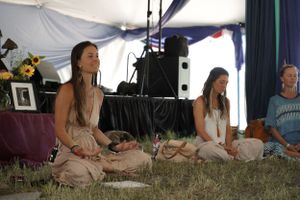 People seated in meditation posture on grass inside a decorated tent space.