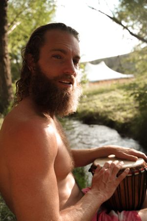 A musician plays a wooden drum in a natural outdoor setting.