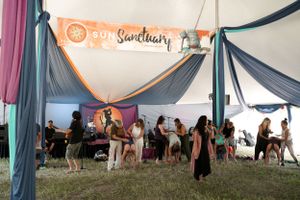 A festival tent with colorful draped fabric and a Sun Sanctuary banner displays people gathering beneath.