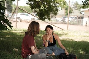 Two people having a conversation while sitting on grass in a shaded outdoor setting.