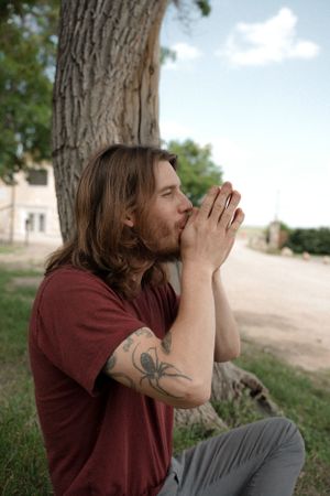 A person with long hair and a tattoo cups their hands around their mouth in an outdoor setting.