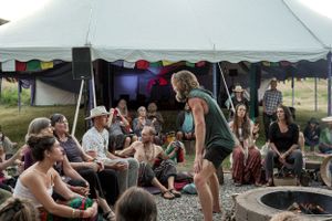 A crowd gathers under a large white tent around a campfire circle.