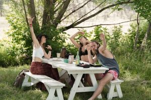 Friends gather around a white picnic table for an outdoor meal under tree branches.