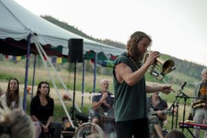 A musician plays trumpet on stage under a white tent while audience members watch.
