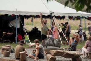 People gather under a white tent for an outdoor community event with log seating.