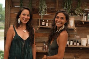 Two women smiling in front of shelves displaying natural products in a rustic wooden setting.