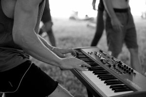 Black and white close-up of hands playing a keyboard synthesizer outdoors.