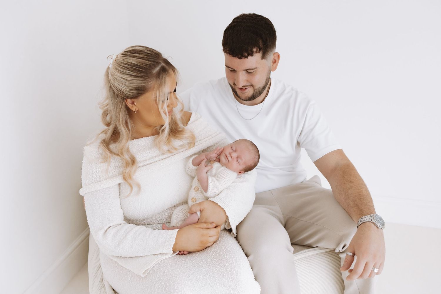 Parents and newborn baby cuddle together on white bedding in tender family moment.