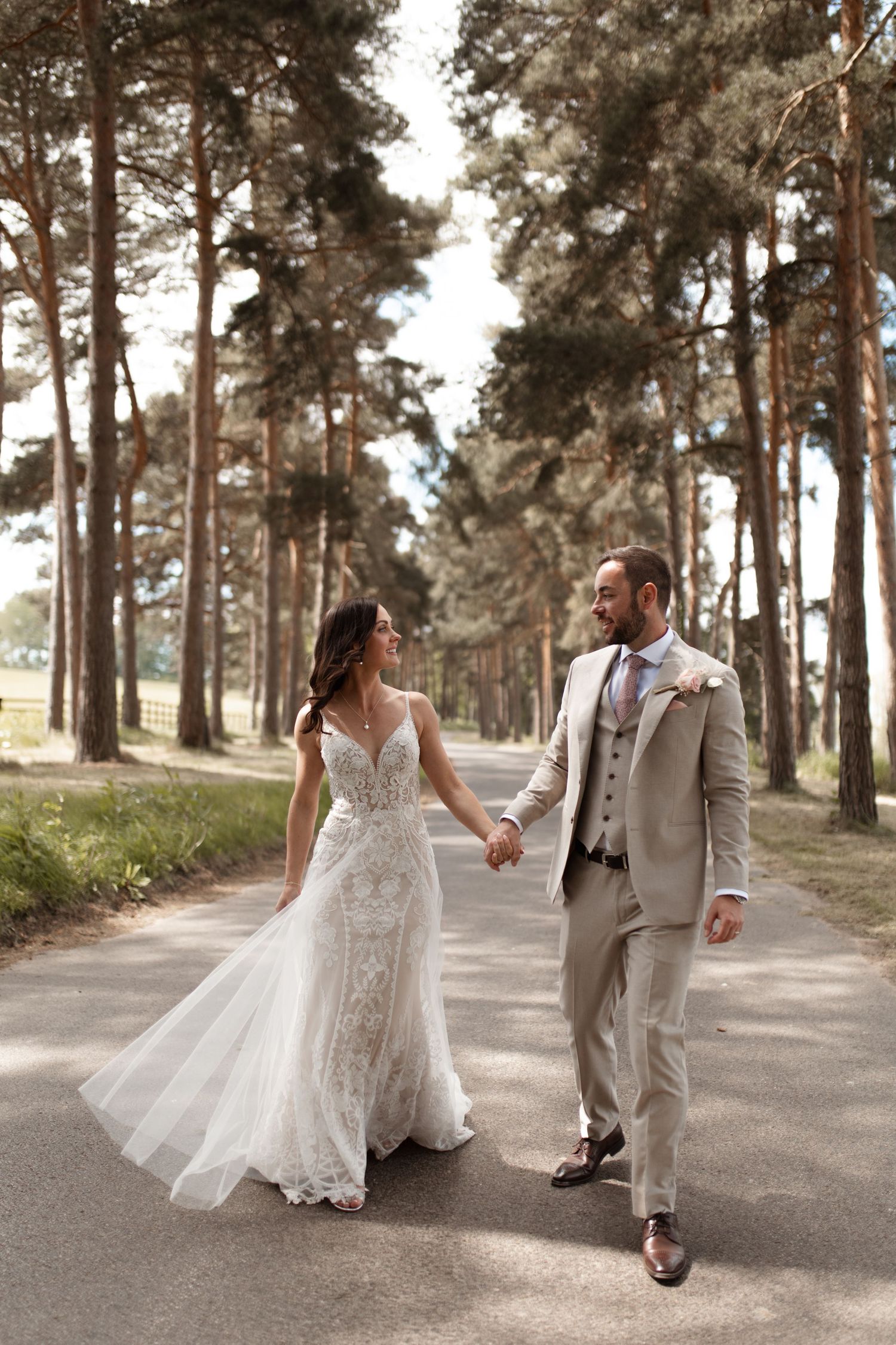 A bride and groom walk hand in hand down a pine tree-lined path during their romantic outdoor wedding photo session.
