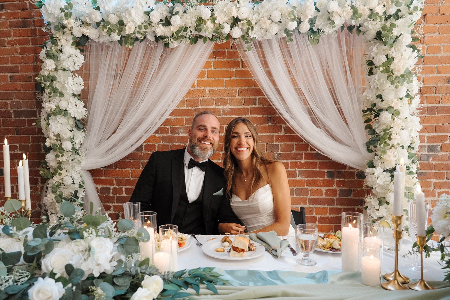 Elegant wedding reception setup with white floral archways, draped fabric, and exposed brick walls.
