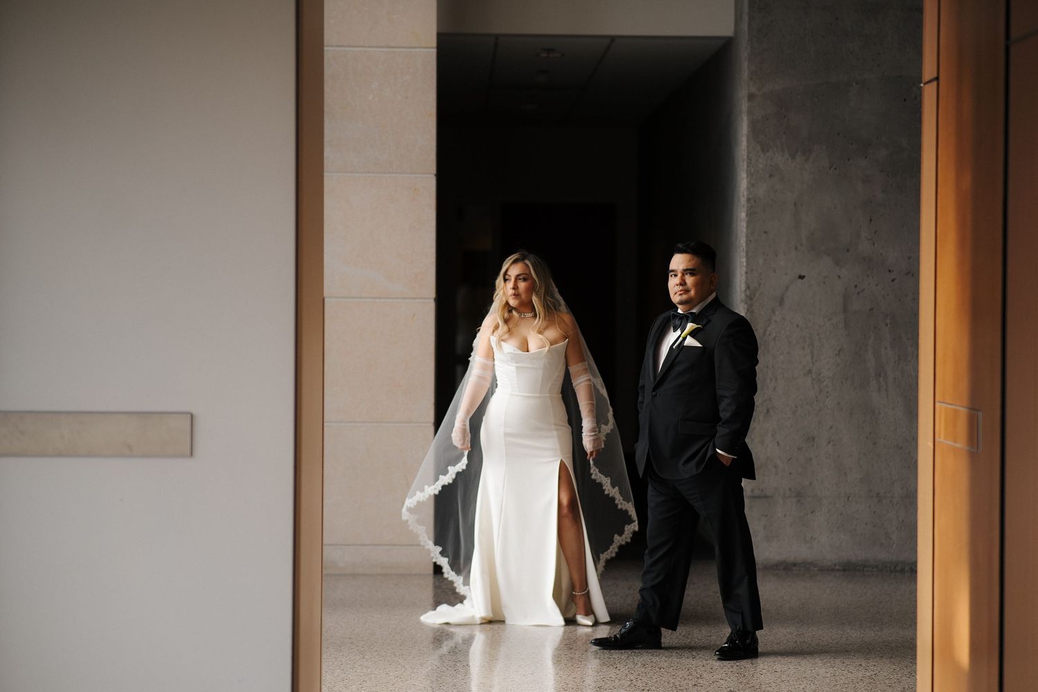 A bride and groom stand together in an elegant minimalist hallway with white walls and wooden accents.