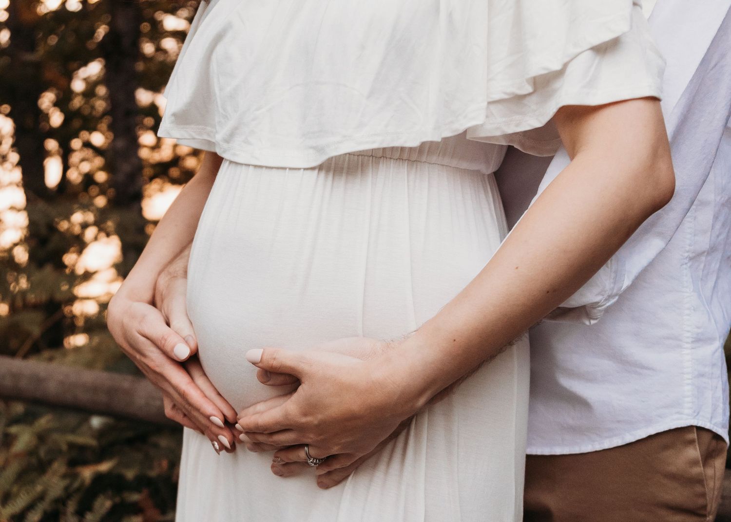 Hands create a heart shape over a pregnant belly in a white maternity dress outdoors at sunset.