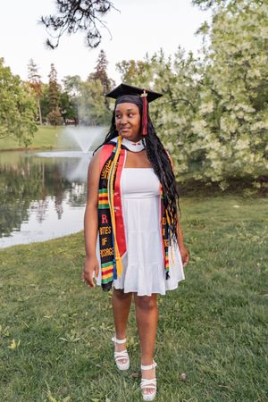 Graduate in white dress and colorful stole poses near a lake during graduation photoshoot.