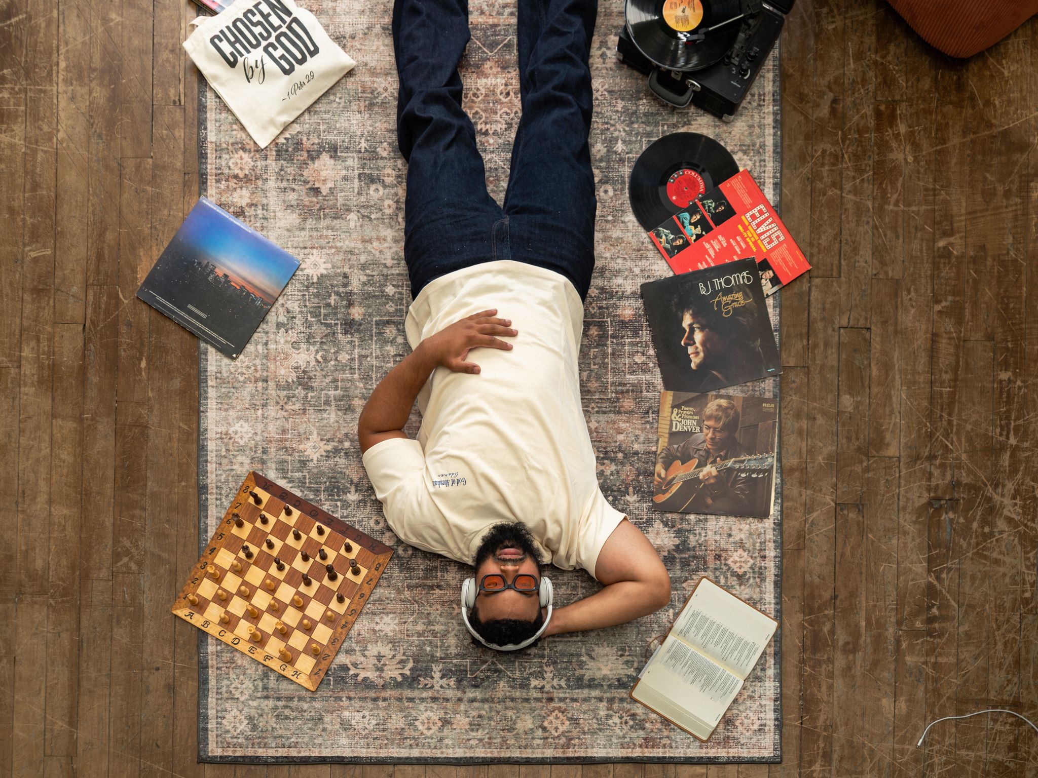 Man lying on rug surrounded by vinyl records, chess board, books, and headphones, viewed from above.