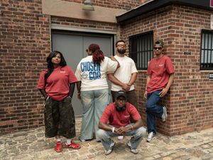 Five young adults posing outdoors against a brick wall, wearing casual streetwear including a Chosen by God graphic tee.
