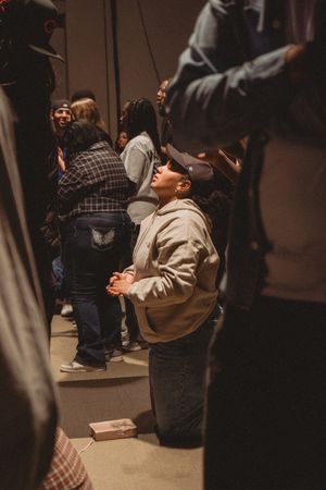 Person kneeling in prayer with hands clasped, looking upward, surrounded by a crowd in a dimly lit gathering.