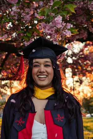Smiling graduate in black cap and gown with red trim poses under blooming pink cherry blossom tree at golden hour.