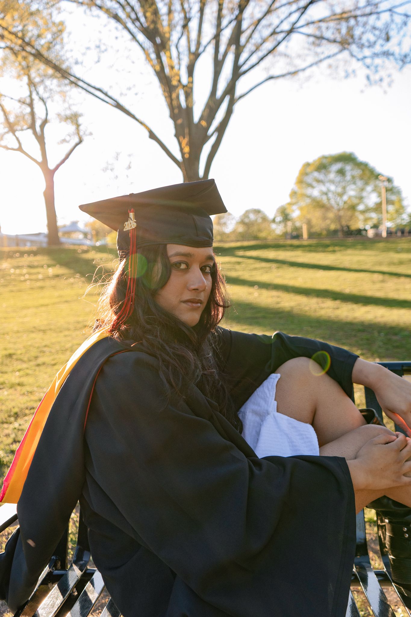Graduate in black cap and gown sits on bench in sunny park, gazing back at camera during golden hour.
